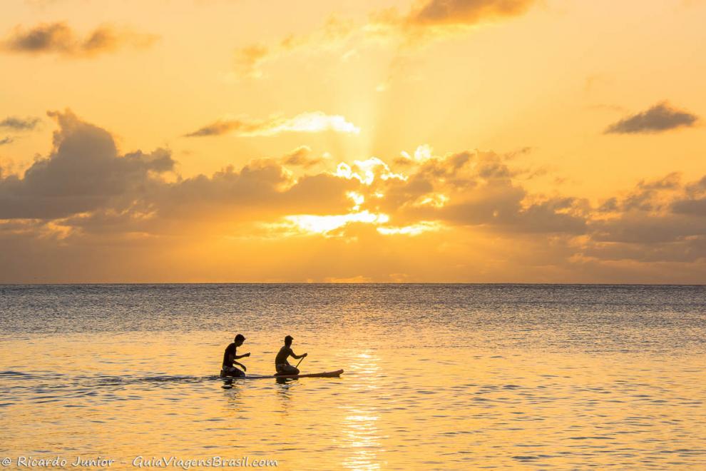 Imagem de duas pessoas remando no mar e ao fundo um céu alaranjado.